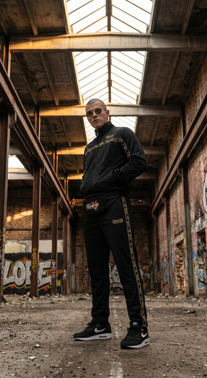 Modello GABBERPLANET indossa tuta 100% Hardcore Essential nera con dettagli dorati in un magazzino industriale